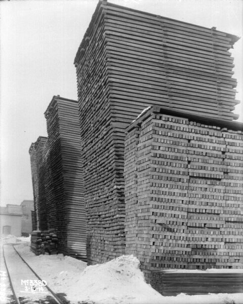 Piles of lumber in plant yard. Snow is on the ground. Railroad tracks are along the left side.