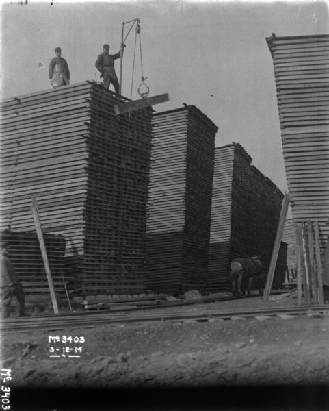Men are standing on top of a pile of lumber unloading pieces of wood with a rope and pulley attached to a pole at the top of the pile. At the base of the lumber piles is a man is standing with a horse who is attached to the other end of the rope. Another man is standing on the far left.
