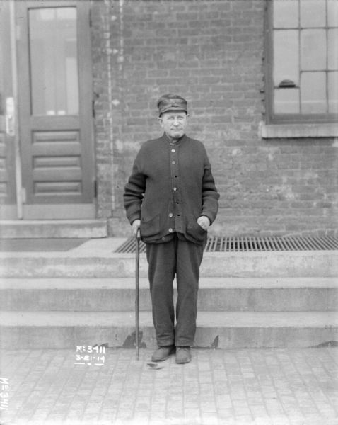 Full-length portrait of a man standing in front of the steps of a brick factory building. He is wearing a hat, sweater and pants, and is holding a walking stick.
