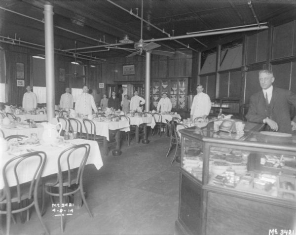 White coated waiters are standing in the dining room interior. A man wearing a suit is standing behind a snack counter in the foreground on the right.