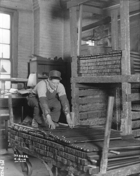 A man is bending over while standing on a wheeled cart and reaching down towards metal bars that are stacked. The forge is to the right of the cart.