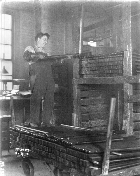 A man is on a wheeled cart on top of a stack of metal bars. The man is holding a metal bar that is resting on another stack of metal bars on a shelf.