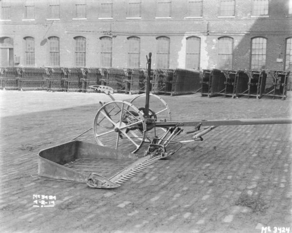 Mower outdoors in the yard at McCormick Works. There are stacks of agricultural parts along a brick wall of a factory building in the background.