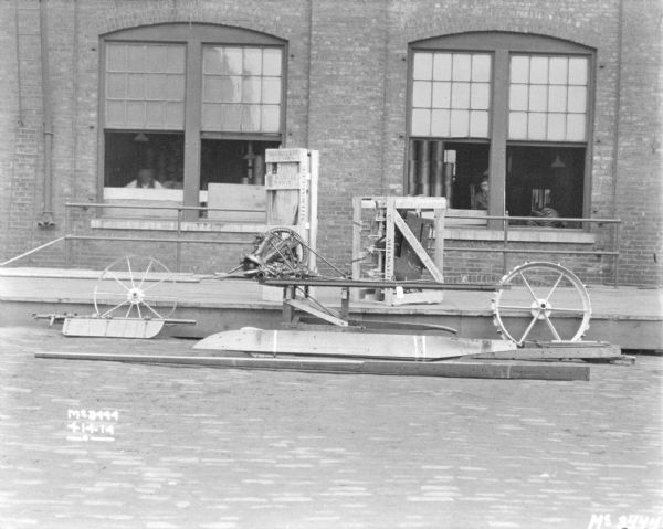 Corn Binder disassembled and banded for shipping outdoors on a loading dock at McCormick Works. People are standing at the windows of the factory in the background.