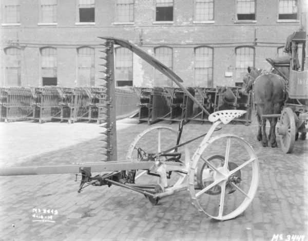 A mower is set up outdoors on the cobblestones in the yard. There are rows of agricultural parts in stacks near the side of a brick factory building in the background. On the far right is a horse-drawn wagon.
