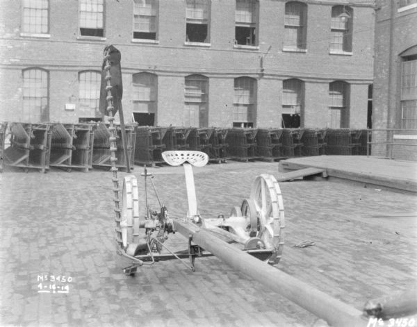 A mower is set up outdoors on the cobblestones in the yard. There are rows of agricultural parts in stacks near the side of a brick factory building in the background. On the far right is a loading dock.