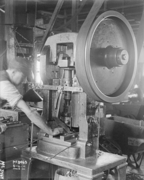 A man wearing a hat is operating a machine that is belt-driven from the ceiling.