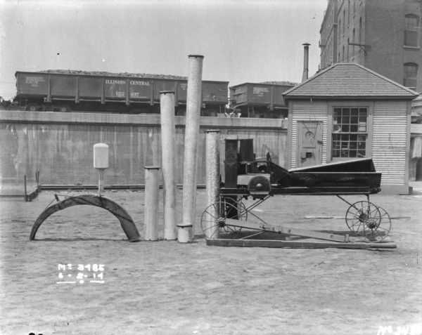 Ensilage cutter, with an assortment of pipes for silo-filling arranged beside it. In the background is a small building near the raised railroad platform. Open railroad cars are on the platform.