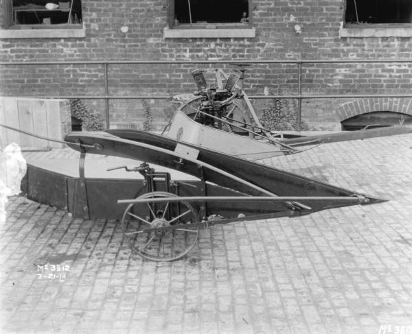 Side view of a Corn Binder outdoors on the cobblestones in the factory yard. A brick factory building is in the background.