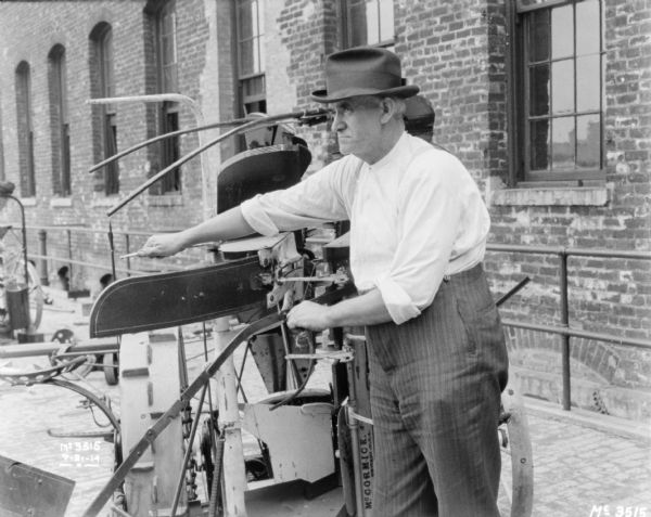 Man demonstrating operation on a Corn Binder outdoors in the factory yard. There is a brick factory building in the background.