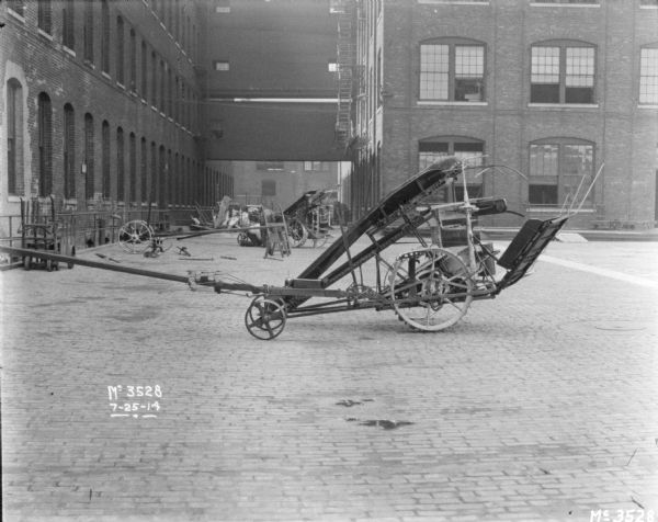 Corn Binder outdoors on the cobblestones in the factory yard cluttered  with machines. Two men are working on a machine in the background. Brick factory buildings are connected by upper level enclosed walkways. 