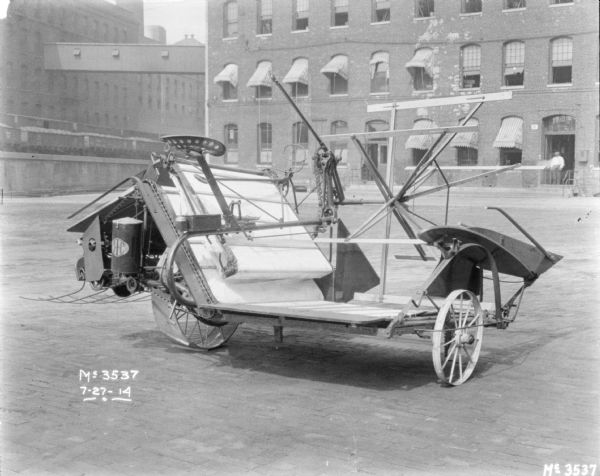 Binder outdoors on the cobblestones in the factory yard. Brick factory buildings and railroad cars on an elevated platform are in the background. A man is standing in an open doorway of the factory.