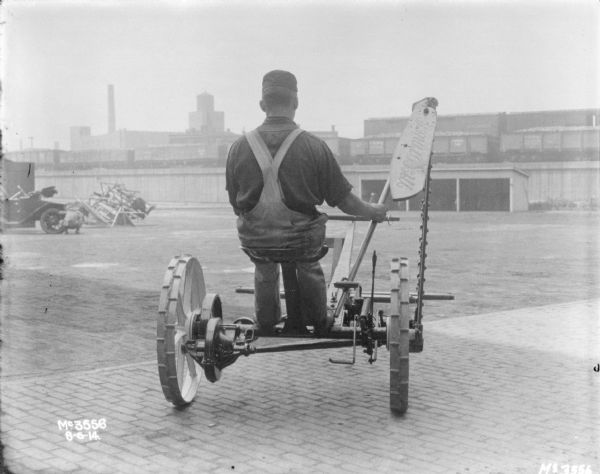 Man riding a McCormick Mower in the factory yard. He is demonstrating the raising of the blade. In the background railroad cars are on an elevated platform, with buildings beyond. On the left a man is looking at the front wheel of an automobile.