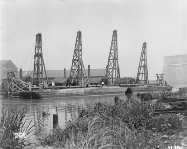 View from shoreline of waterfront with docks and cranes, and factory buildings behind. Men are standing on a long barge near the cranes.