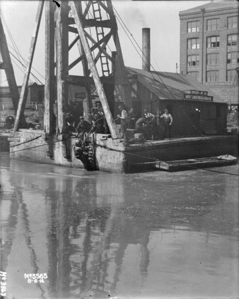 View across water towards a man in a diving suit on a ladder that is just above the water. A group of men are standing on the dock nearby. A sign on the building behind the crane reads: "Great Lakes Dredge and Dock Co., Driver No. 12."