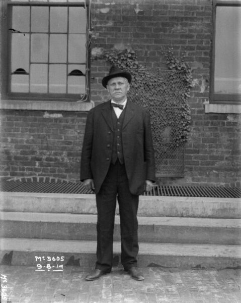 A man wearing a suit is standing outdoors at McCormick Works. A brick factory building is behind him.