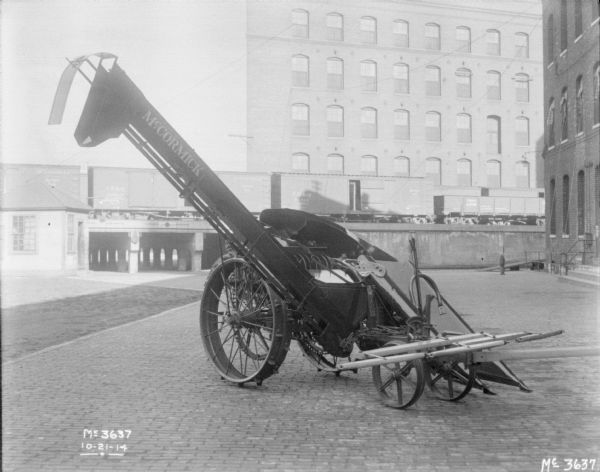 Corn Picker outdoors on the cobblestones in the factory yard. In the background are railroad cars are on an elevated platform, and a large brick building.