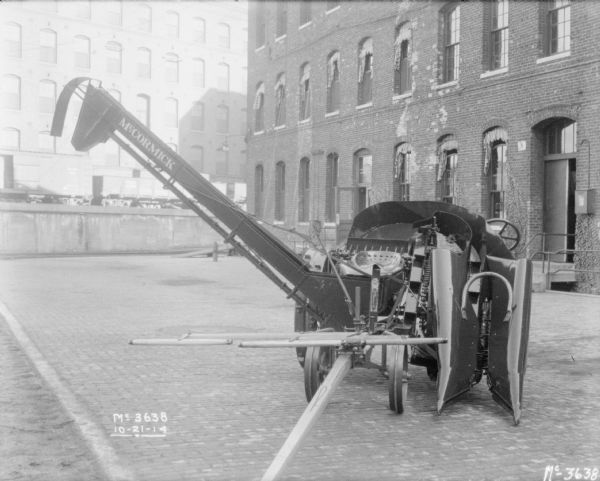 Corn Picker outdoors on the cobblestones in the factory yard. There is a brick factory building on the right, and in the background are railroad cars are on an elevated platform, and a large brick building. 
