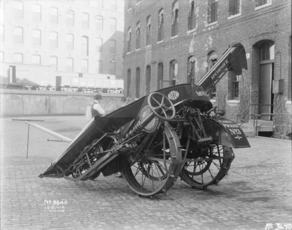 Corn Picker No. 3 outdoors on the cobblestones in the factory yard. A man is standing behind the picker. There is a brick factory building on the right, and in the background are railroad cars are on an elevated platform, and a large brick building. 