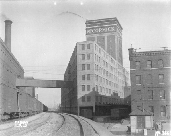 View from elevated railroad tracks towards railroad yard, and newly constructed plant addition. Men are standing in the yard below. A tower on one of the buildings has a sign near the roof that reads: "McCormick." A man standing on the roof above appears to be spraying water into the air from a hose.