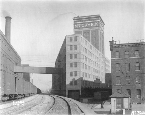 View from elevated railroad tracks towards railroad yard, and newly constructed plant addition. Men are in the yard below. A tower on one of the buildings has a sign near the roof that reads: "McCormick." Two men standing on the roof above appear to be spraying water into the air from a hose.