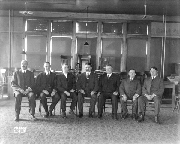 Six men in business suits are sitting in a row on chairs in a room. The shades on the windows in the background have been closed, and there are chairs and other furniture stacked up behind the group of men.