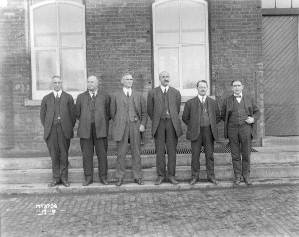 Outdoor group portrait of six men wearing business suits standing outdoors in front of a brick factory building.