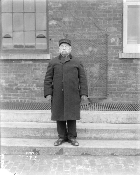 A man is standing in front of a brick factory building. He is wearing a coat and a hat.