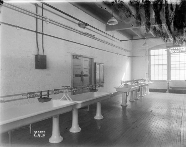 View of a large employee washroom, with large sinks with pedestal bases on a hardwood floor. Large windows are on the wall in the background, and the ceiling rafter's are exposed.