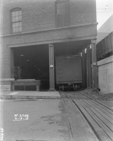 Boxcar entering plant yard. A man is standing on the loading dock which is under the factory building. On the right alongside the building are railroad cars on an elevated platform.