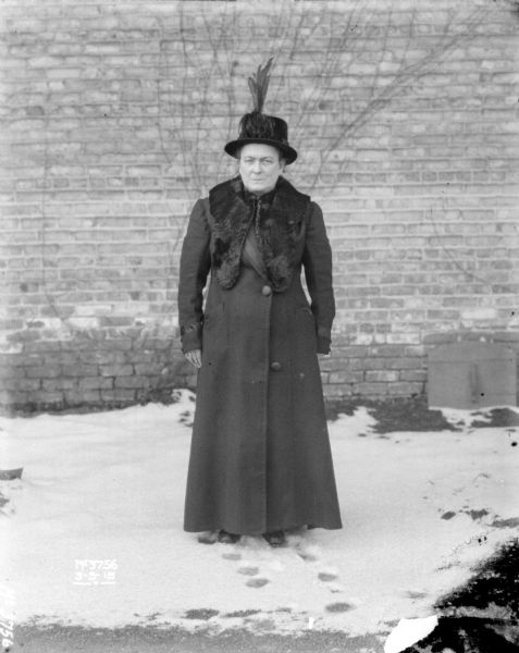 Woman posing outdoor in the snow. She is wearing a full-length coat with a fur collar, and a hat with tall feathers. A brick factory building is in the background.