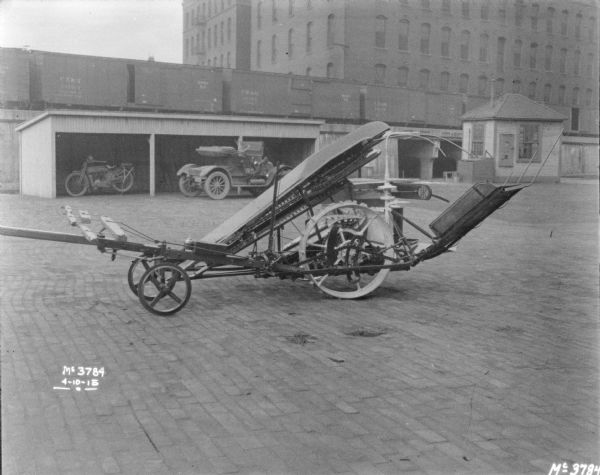 Close-up of Corn Binder set-up on cobblestones in the factory yard. In the background a motorcycle is parked underneath an open-sided wood shed. In front of the shed a man is near an automobile. Behind the shed are railroad cars on an elevated platform. A small building, perhaps a gatehouse, is near the underpass of the railroad platform.