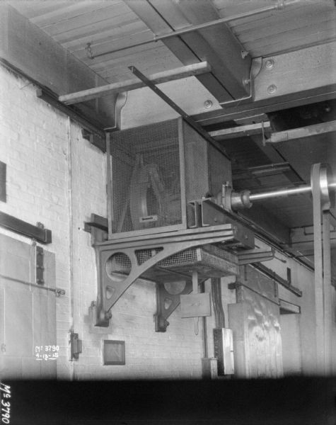 View from left of machinery mounted on brick wall at the ceiling beams, which is powering a wheel on an axle which is spinning a belt that is attached to something (out of frame) on the floor. The tops of large barn-style doors are on the left and right of the machinery. There is a protective screen around and underneath the machinery.