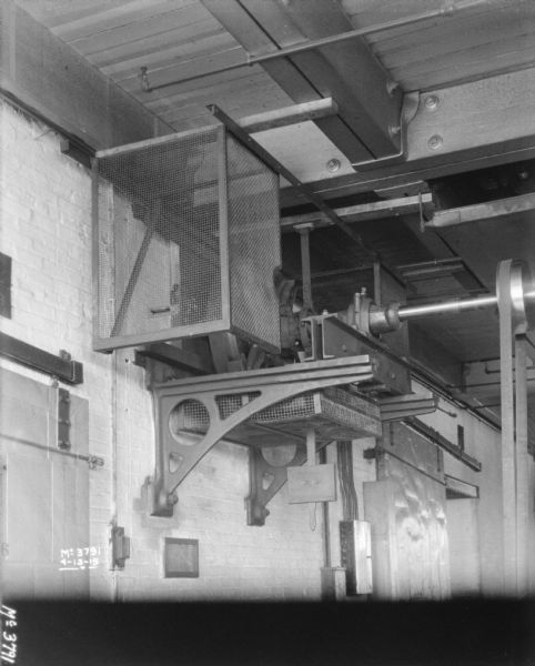 View from left of machinery mounted on brick wall at the ceiling beams, which is powering a wheel on an axle which is spinning a belt that is attached to something (out of frame) on the floor. The tops of large barn-style doors are on the left and right of the machinery. The protective screen on the left side has been pulled out and away from the center axle.