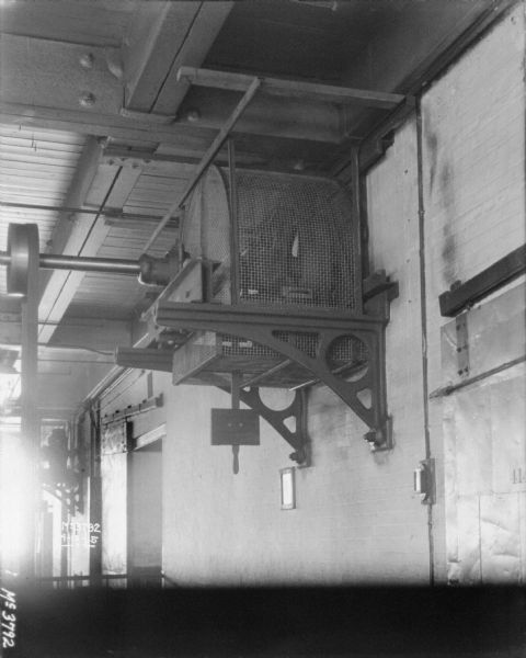 View from right of machinery mounted on a brick wall up at the ceiling beams, which is powering a wheel on an axle which is spinning a belt that is attached to something (out of frame) on the floor. The tops of large barn-style doors are on the left and right of the machinery. There is a protective screen around and underneath the machinery.