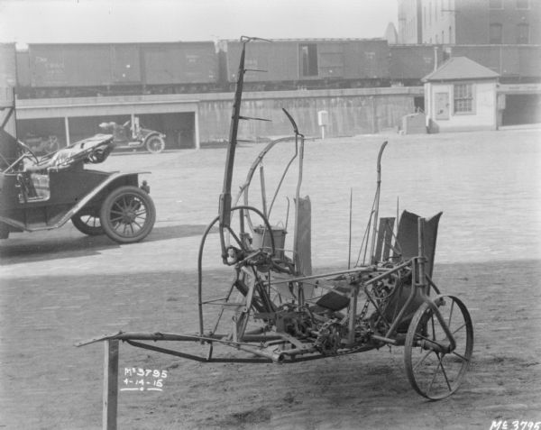 A piece of machinery is set-up on the cobblestones. There is an automobile parked on the left. In the background is a motorcycle parked in an open-sided shed, with another automobile parked in front, and a small, wooden-sided building, perhaps a gatehouse, which is near the underpass of an elevated platform that railroad cars are sitting on. In the far background are buildings.