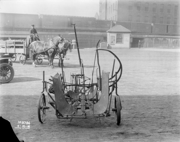 A piece of machinery is set-up on the cobblestones. There is an automobile parked on the left, and behind it is a man sitting on a horse-drawn wagon. In the background is a small, wooden-sided building, perhaps a gatehouse, which is near the underpass of an elevated platform that railroad cars are sitting on. In the far background are buildings.