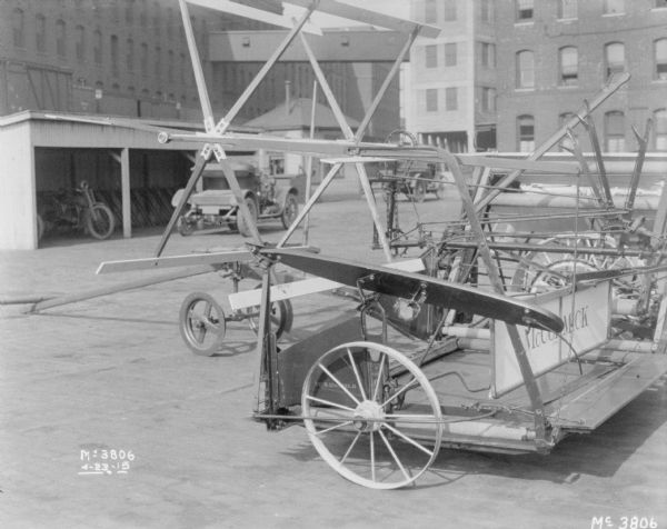 McCormick Binder set-up outdoors in the factory yard. In the background is a motorcycle parked in an open-sided shed, with another automobile parked in front, and a small, wooden-sided building, perhaps a gatehouse, which is near the underpass of an elevated platform that railroad cars are sitting on. In the far background are buildings.