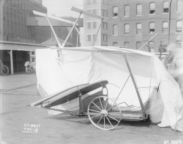 A McCormick Binder is set-up outdoors on the cobblestones in the factory yard. A white cloth has been draped over the binder, showing the side. In the background is an automobile parked near an open-sided shed, and brick factory buildings, with a skybridge between two of the buildings.