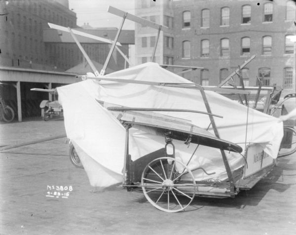 Binder set-up outdoors in factory yard, with cloth masks to accentuate parts. In the background is a motorcycle parked in an open-sided shed, with another automobile parked in front. In the far background are buildings.