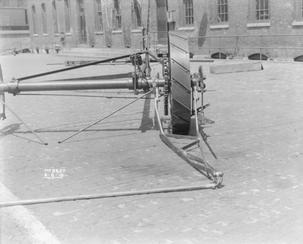 Close-up of wheels on a binder set-up outdoors in the factory yard. A brick building is in the background, and behind it are railroad cars on an elevated platform.