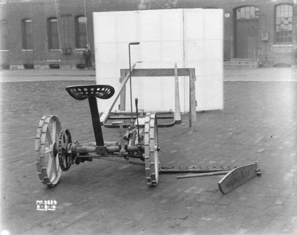 Rear view of a mower outdoors in the factory yard. A small backdrop is set up behind the mower. In the background a man is standing in front of a brick factory building.