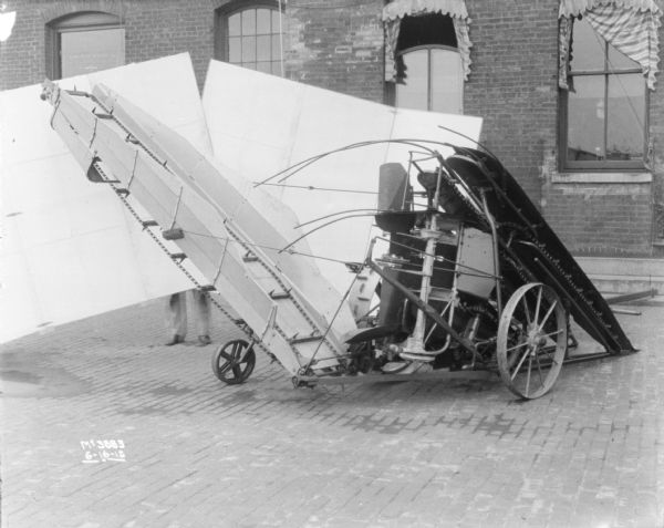Corn Binder set-up on the cobblestones in factory yard. Men (obscured) are holding a backdrop set-up behind the mower. In the background is a brick factory building.