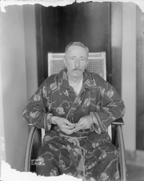 Portrait of a man sitting in a wheelchair wearing a bathrobe, holding reading glasses.