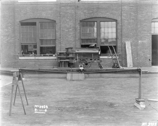 Evener set up on sawhorses outdoors in factory yard. Printed on the evener: "New McCormick Self Dump Rake." A brick factory building and a loading dock are in the background. The large windows are open.