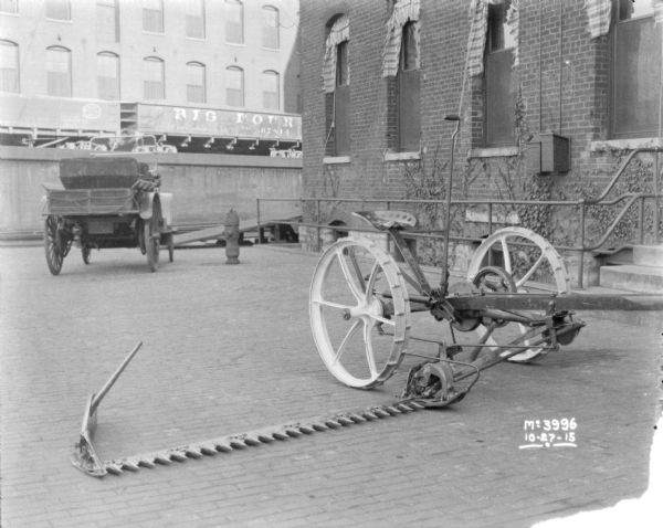 Mower displayed outdoors in the yard at McCormick Works. A brick factory building is on the right, and an open automobile or truck is parked near a ramp. In the background railroad cars are on an elevated platform.