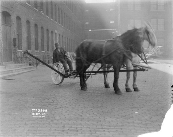 A man is driving a horse-drawn mower in the yard at McCormick Works. Brick factory buildings are in the background.