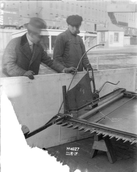 Two men examining binder labeled parts outdoors in the factory yard. Brick factory buildings and railroad cars on an elevated platform are in the background.