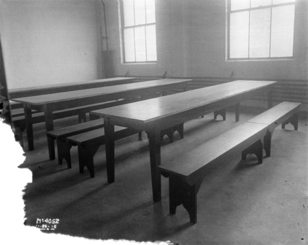 Long tables and benches in the dining room. Large windows are on the wall in the background.