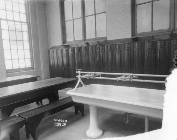 Lavatory interior with a long sink in the foreground,  and lockers underneath a window behind. On the left are long tables and benches, and a large window along the brick wall.